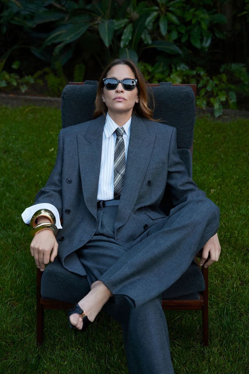 Woman in an oversized grey suit sits in a chair in a garden setting, wearing sunglasses and gold bracelets. Editorial fashion.