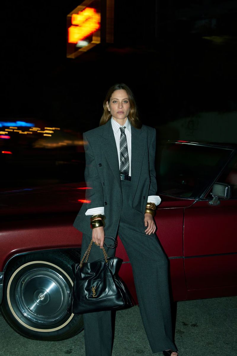 Woman in grey suit poses next to a classic car at night, accessorized with gold bangles and a black YSL bag, under the glow of a sign.
