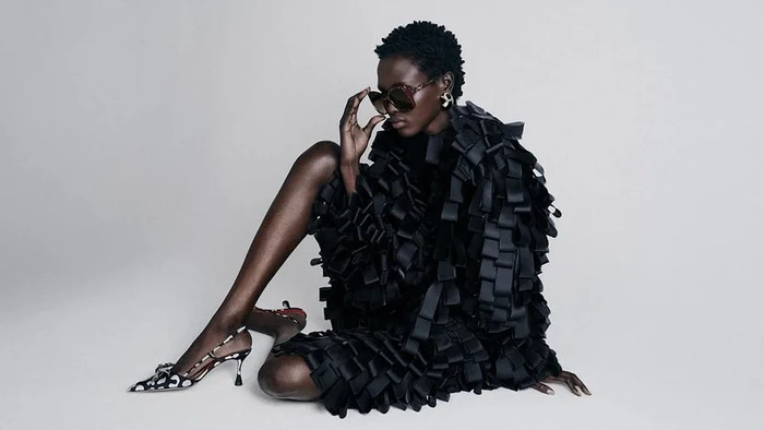 Model Amar Akway in oversized sunglasses wearing a black textured coat and patterned slingback shoes seated on a white floor