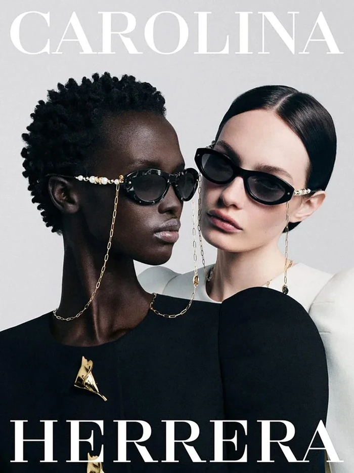 Two female models wearing designer sunglasses with gold chains against a plain light background for Carolina Herrera Eyewear.