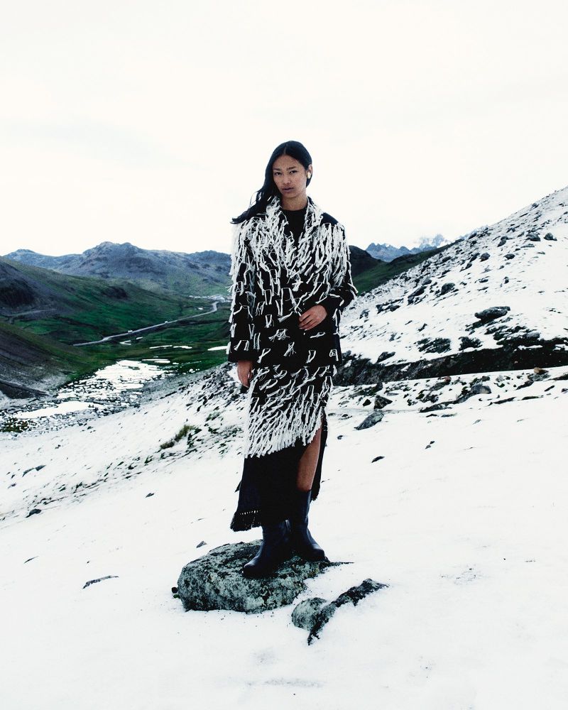 Asian woman stands on a rock in a snowy mountain landscape wearing a black and white fringed coat and black boots on an overcast day.
