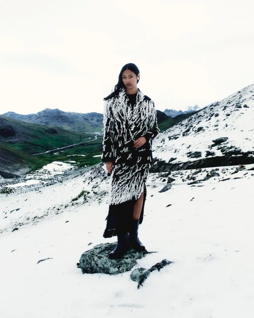 Asian woman stands on a rock in a snowy mountain landscape wearing a black and white fringed coat and black boots on an overcast day.