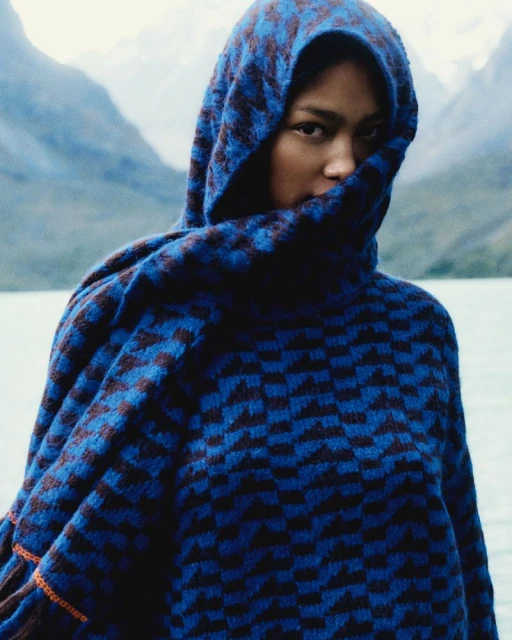 Woman wearing a patterned blue hooded garment posing in front of a mountain lake. The natural lighting gives the scene a soft glow.