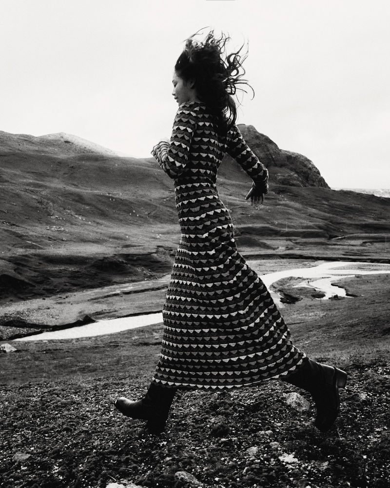 A woman in a patterned dress runs across a rocky landscape, with rolling hills in the background in this black and white outdoor shot.