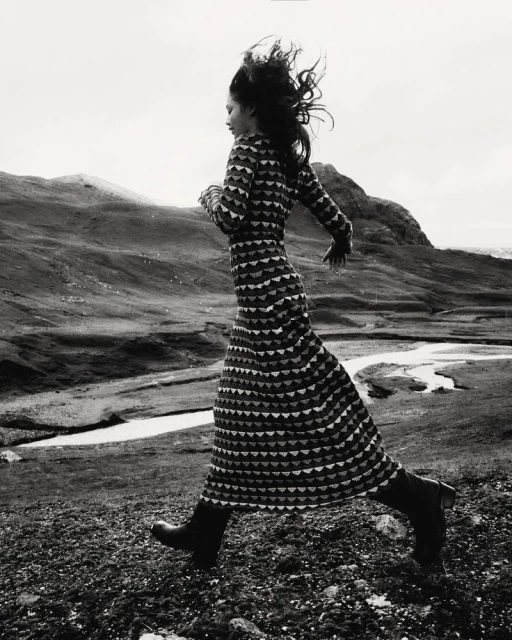 A woman in a patterned dress runs across a rocky landscape, with rolling hills in the background in this black and white outdoor shot.