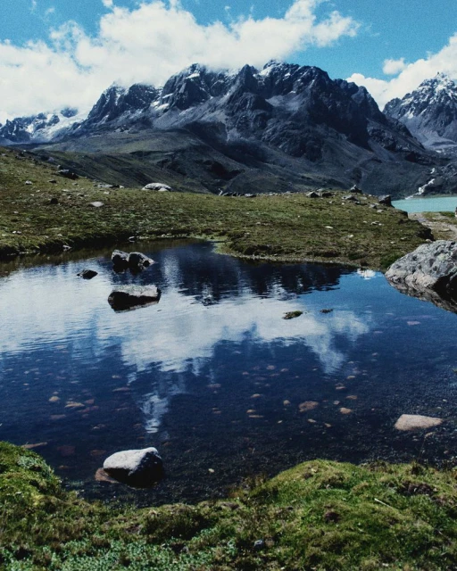 Mountain range and small lake reflecting the sky and clouds, creating a scenic view of nature with rugged peaks and green vegetation.