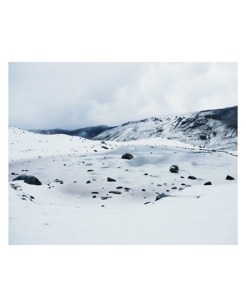 Snow-covered landscape with low mountains and scattered rocks under an overcast sky, creating a serene winter scene.