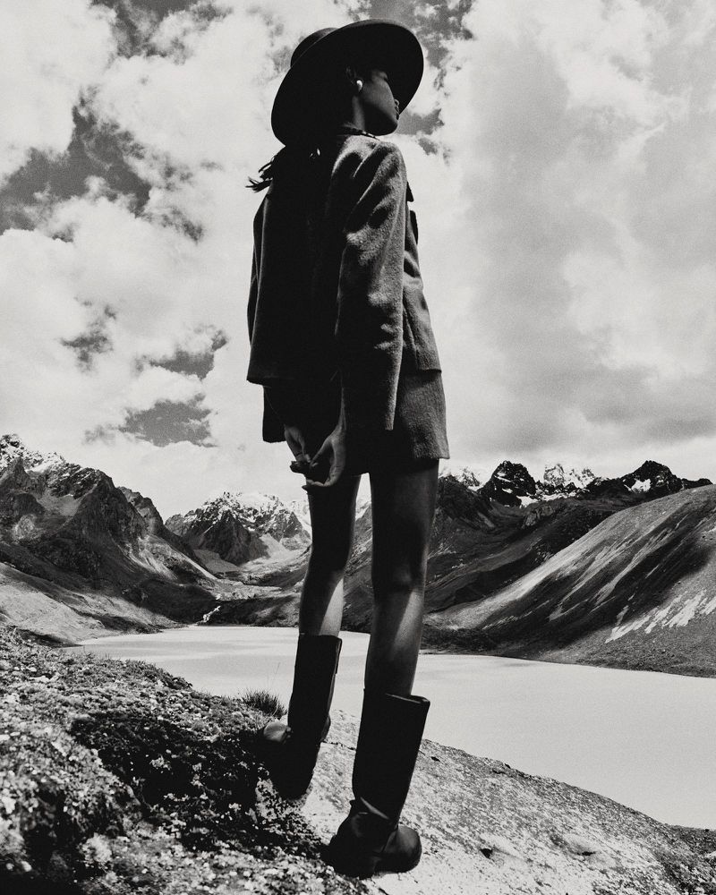 Model standing on a rocky mountain ridge wearing a wide-brimmed hat, jacket, and tall boots against a dramatic mountain and lake background.