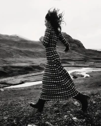Model in patterned long sleeve dress running through a rugged landscape with hills and a river