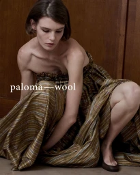 Model crouching against a wooden wall wearing a gathered olive plaid strapless dress for Paloma Wool SS27.
