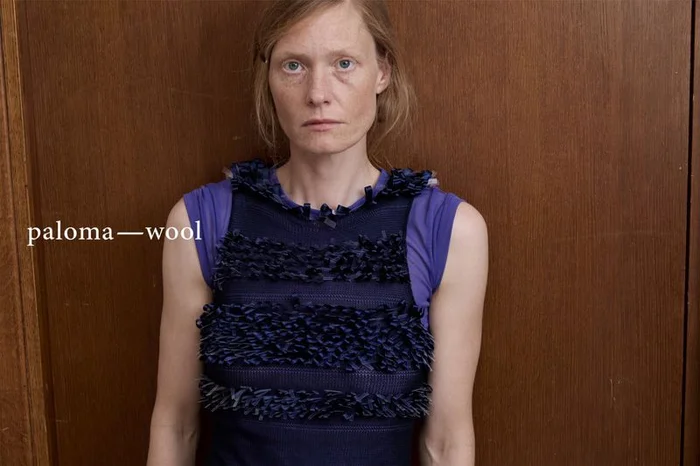 Portrait of a woman wearing a textured navy blue top over a purple t-shirt against a wooden wall.