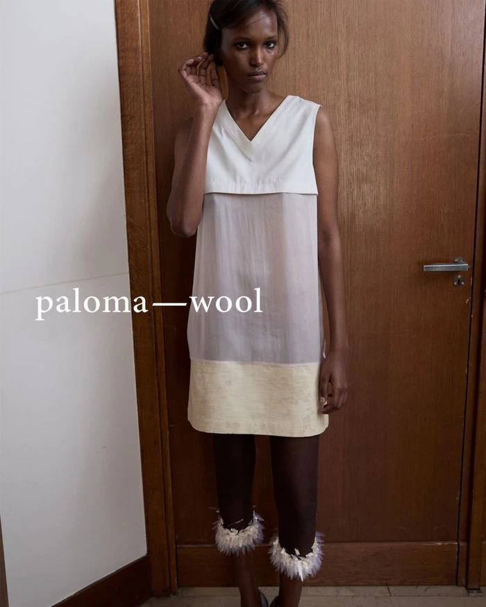 Model wearing a layered tunic dress and feather-trimmed leggings standing before a wooden door.