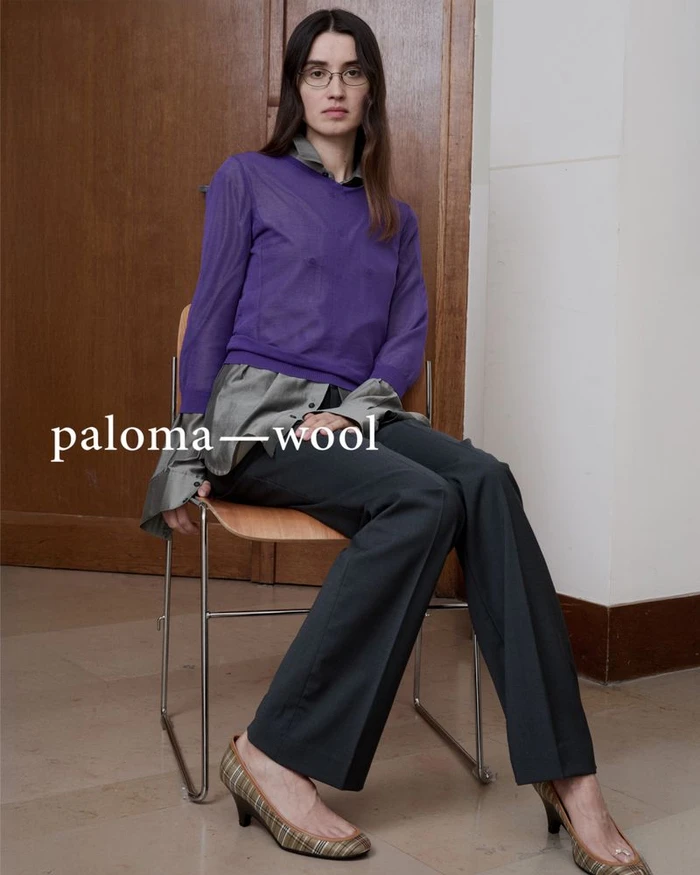 Woman seated on a metal chair wearing a purple top over a grey shirt, dark trousers, and plaid heels against a wood door.