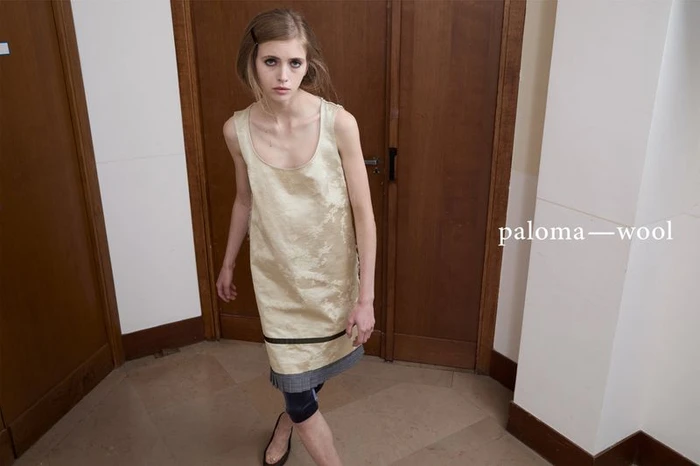 Model wearing a textured cream dress standing in a hallway with wooden double doors for Paloma Wool