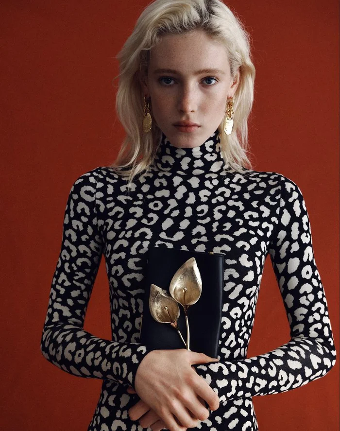 Model with blonde hair wearing a black and white leopard print turtleneck holding a black clutch with gold floral hardware
