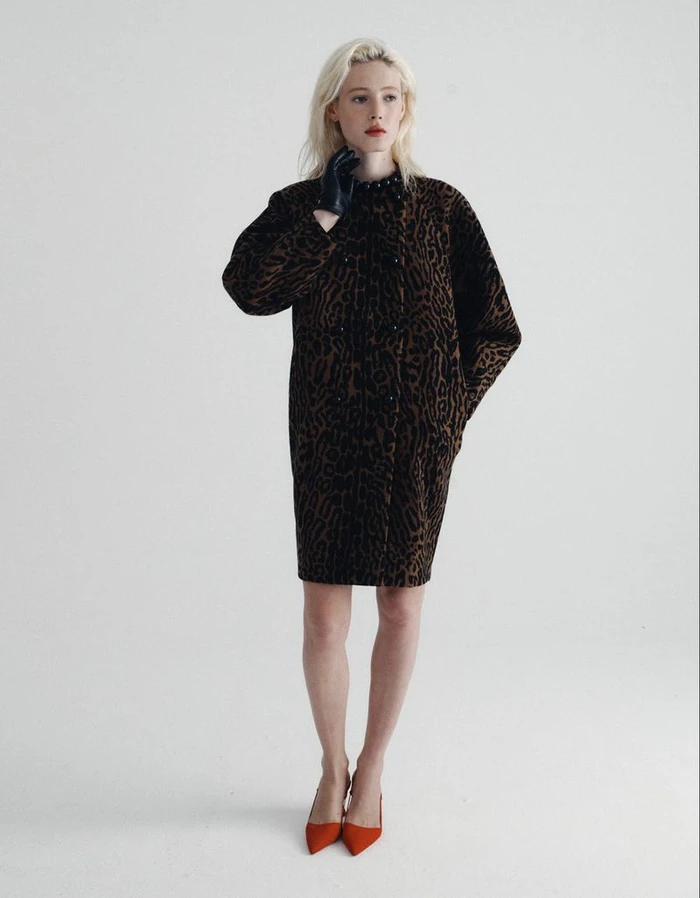 Model with blonde hair wearing an animal-print coat and red heels standing against a seamless white background.
