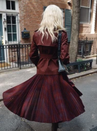 Model with long blonde hair wearing a burgundy trench coat and pleated skirt walking on a city street.