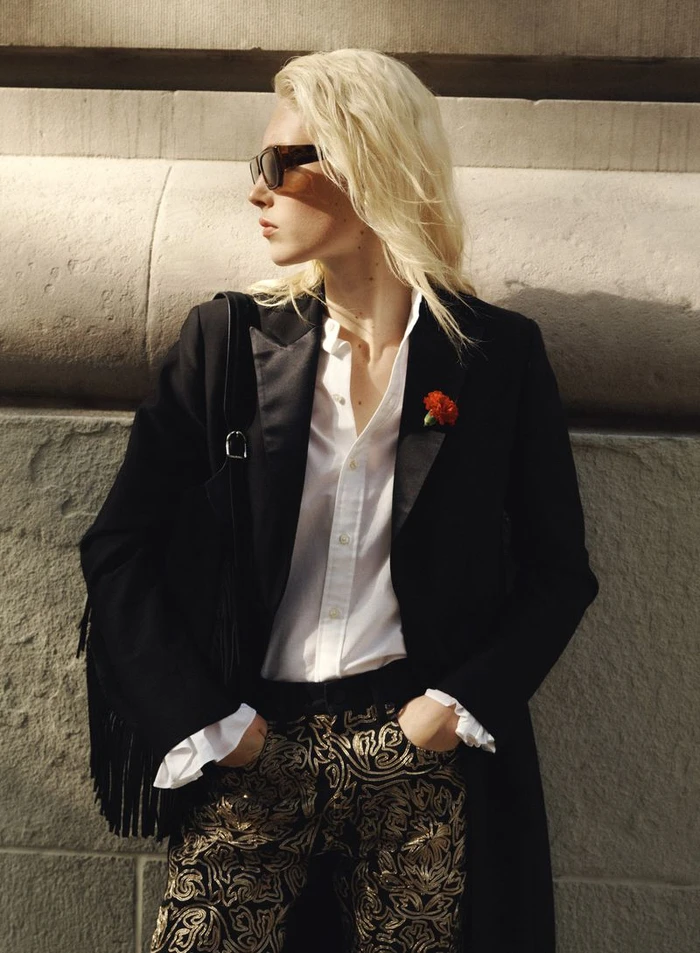 Model Summer Dirx wearing a black coat, white shirt, and patterned pants against a stone wall