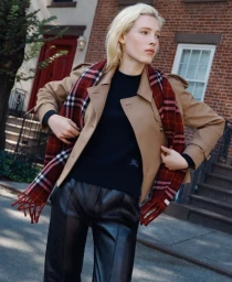 Blonde model wearing a camel trench coat, black top, leather trousers, and a red plaid scarf outdoors on a city street.