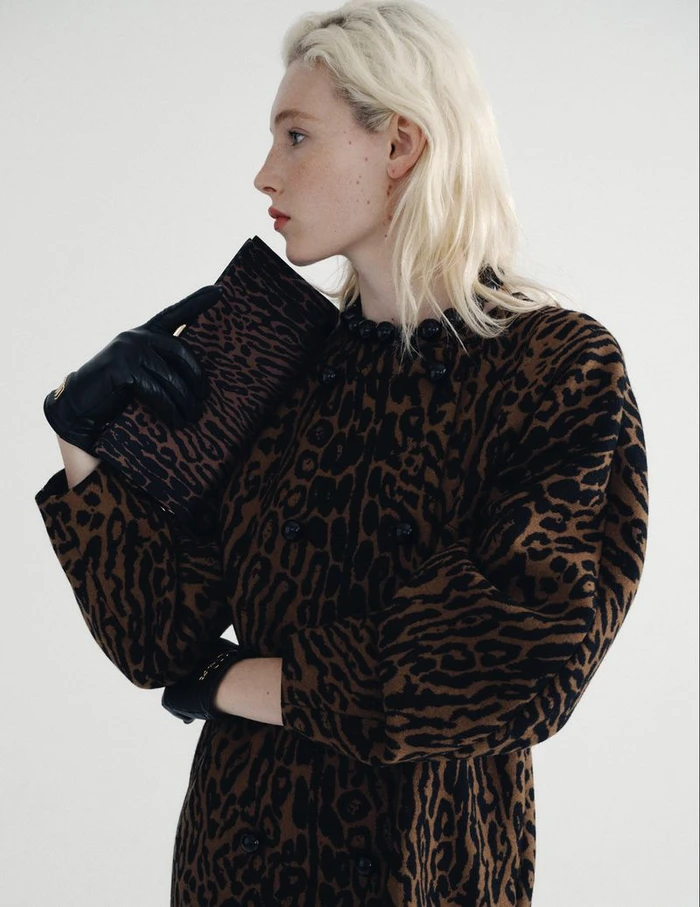 Model with blonde hair wearing a leopard print dress and black leather gloves holding a matching clutch bag