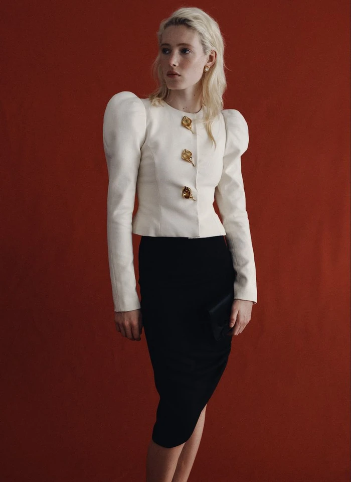 Model Summer Dirx wearing a white structured puff-shoulder top and black skirt against a rust red backdrop for Saks.