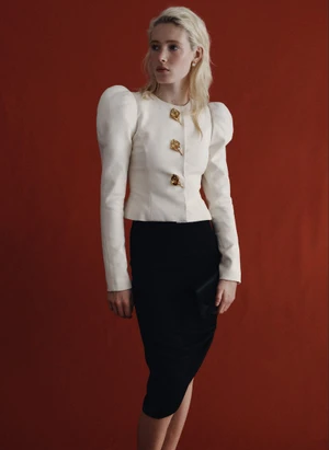 Model Summer Dirx wearing a white structured puff-shoulder top and black skirt against a rust red backdrop for Saks.