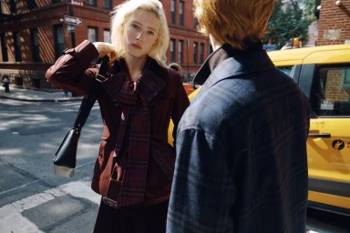 Blonde female model in a burgundy trench coat and plaid bow tie standing on a city street near a yellow taxi cab