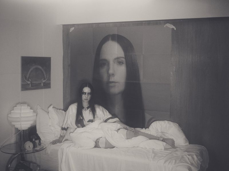 Monochrome portrait of a young woman in bed with striking makeup, alongside a large poster of a woman's face, evoking an intimate mood.