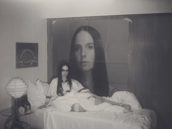 Monochrome portrait of a young woman in bed with striking makeup, alongside a large poster of a woman's face, evoking an intimate mood.