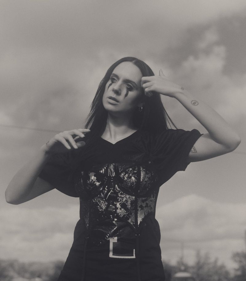 Artistic portrait of a woman with black eye makeup wearing a corset and t-shirt, posing outdoors against a cloudy sky.