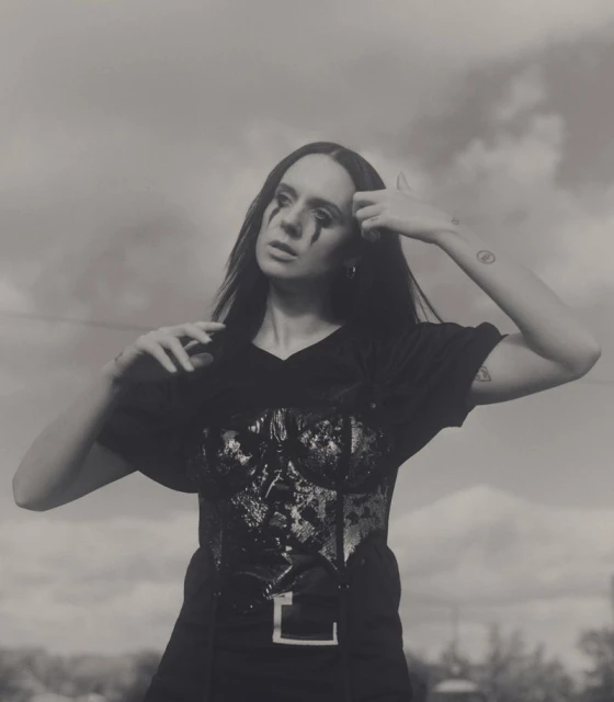 Artistic portrait of a woman with black eye makeup wearing a corset and t-shirt, posing outdoors against a cloudy sky.