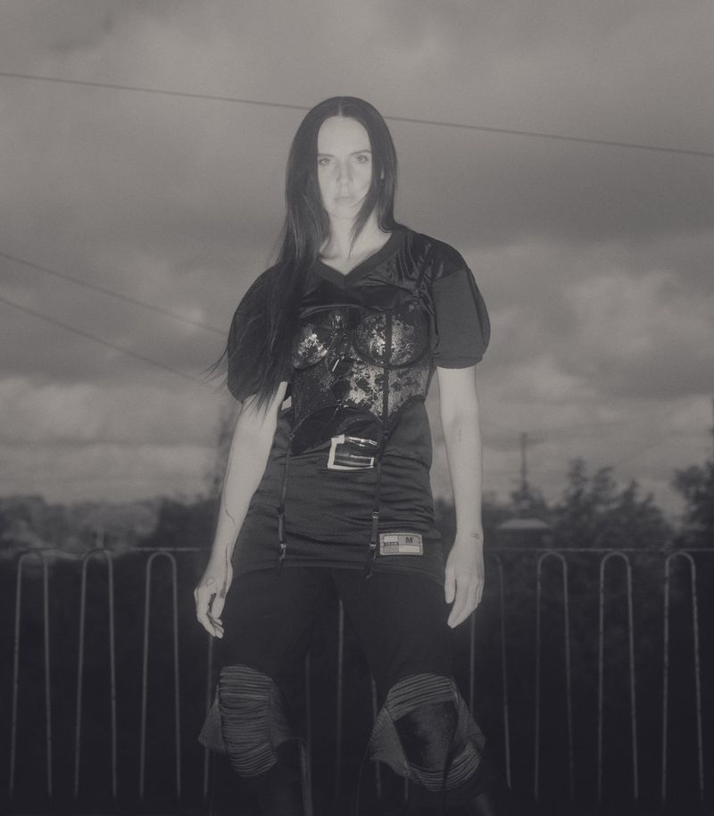 A woman in black athletic wear stands outdoors against a cloudy sky. She is wearing protective pads on her knees.