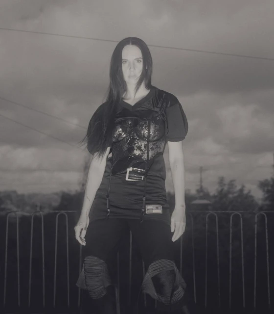A woman in black athletic wear stands outdoors against a cloudy sky. She is wearing protective pads on her knees.