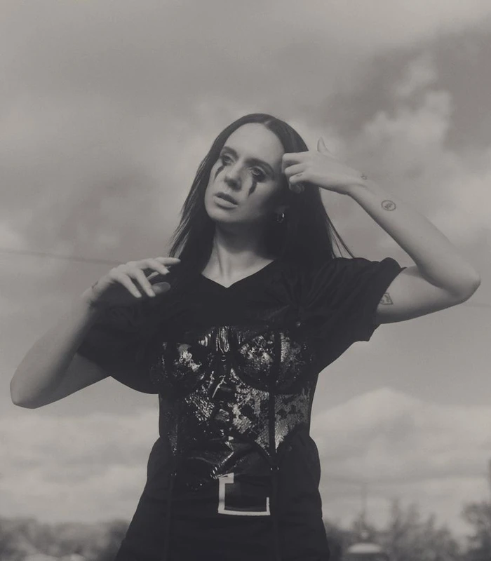 Singer MØ with running black eye makeup wearing a sequin corset and black t-shirt outdoors against an overcast sky.
