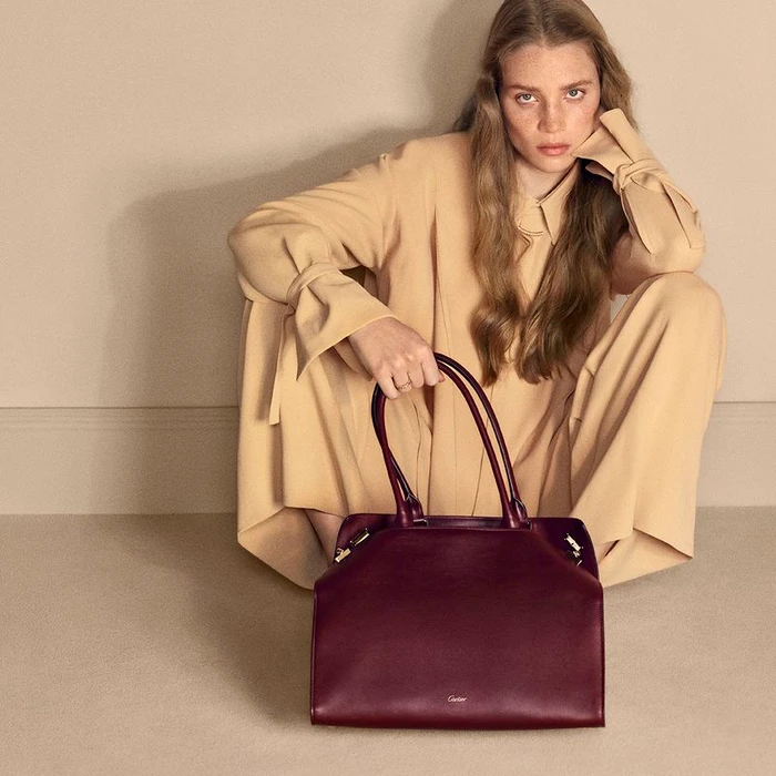Model Rebecca Leigh Longendyke in a beige outfit holding a burgundy leather tote bag for Cartier by Mark Kean.