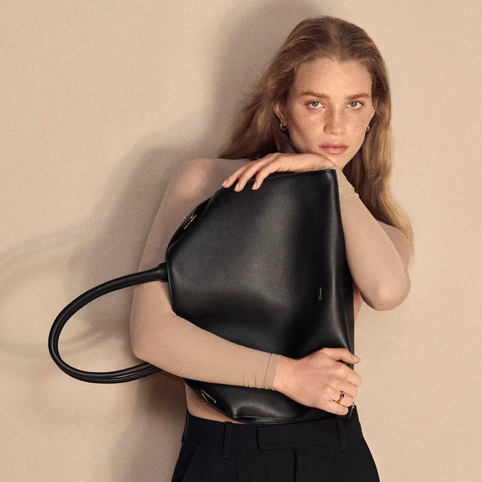 Model Rebecca Leigh Longendyke holding a black leather tote bag against a neutral background for Cartier.