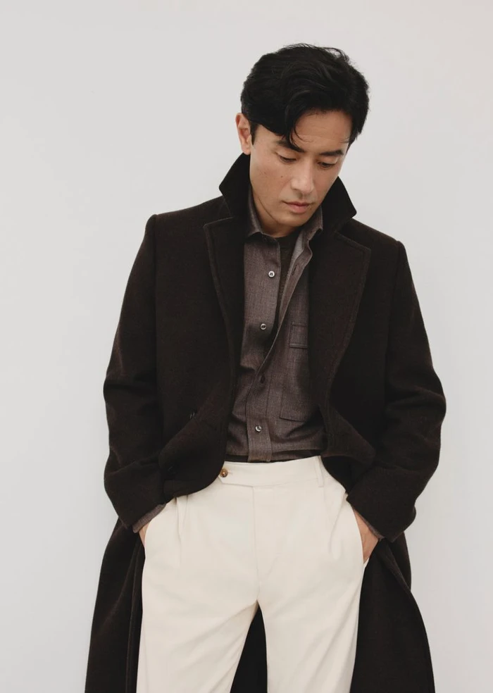 Man wearing a dark coat and cream trousers with his hands in his pockets against a white backdrop.