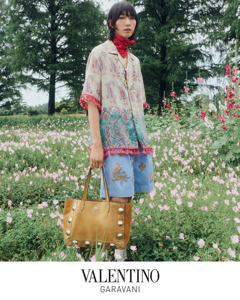 A person stands in a field of flowers, wearing a patterned shirt and shorts, holding a studded tote bag, with trees in the background.