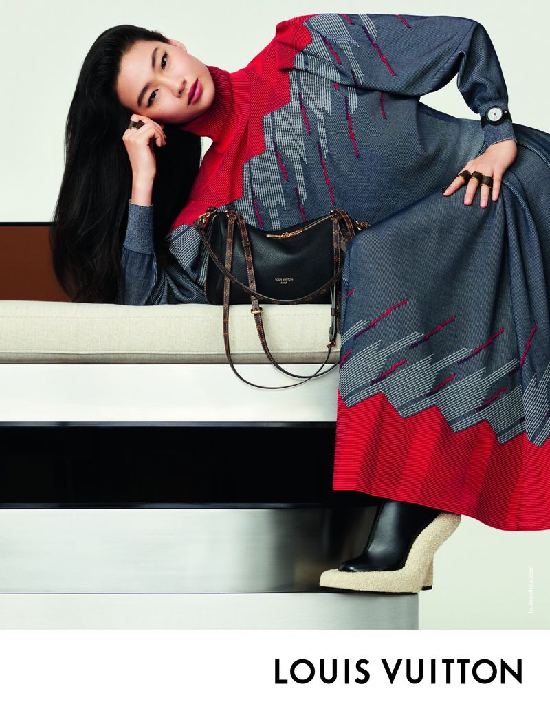 Asian woman reclining in fashionable red and grey patterned dress next to a black purse and wearing black platform heel boots.