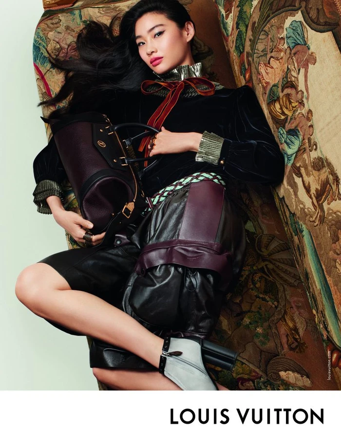 Model lying on an ornate patterned sofa wearing a dark velvet top and holding a dark leather handbag.