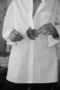 Model wearing a white button-up shirt holding a dried leaf in both hands against an out-of-focus background.