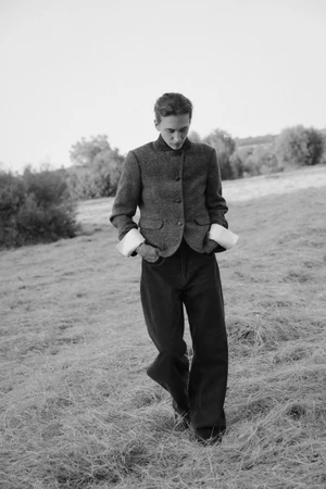 Model wearing dark tweed jacket and trousers walking across a grassy field under an overcast sky.