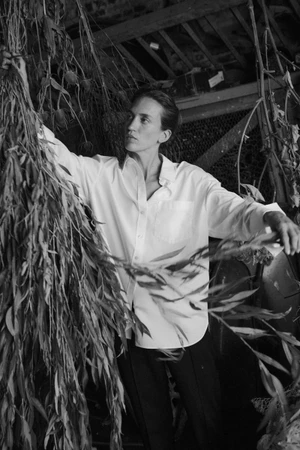 Model in white button-down shirt reaching for hanging dried branches in a rustic interior setting