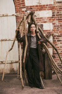 Model standing beneath a wooden branch structure wearing a long brown leather coat against a brick wall background