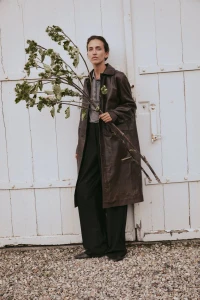 Model wearing a dark leather coat holding tall leafy branches against large white wooden doors.