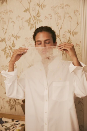 Model holding a translucent patterned fabric across her face, wearing a white button-down shirt against floral wallpaper.