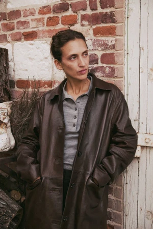 Model wearing a dark brown leather coat and grey polo shirt standing against a weathered brick and white door exterior wall