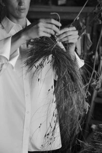 Close-up monochrome image of a woman tying a bundle of dried grass with string, wearing a white button-down shirt.