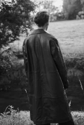 Model seen from behind wearing a long dark leather coat outdoors near grassy fields and water.