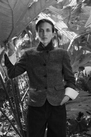 Model wearing a dark textured button-up jacket and dark trousers framed by large tropical foliage in a black and white portrait.